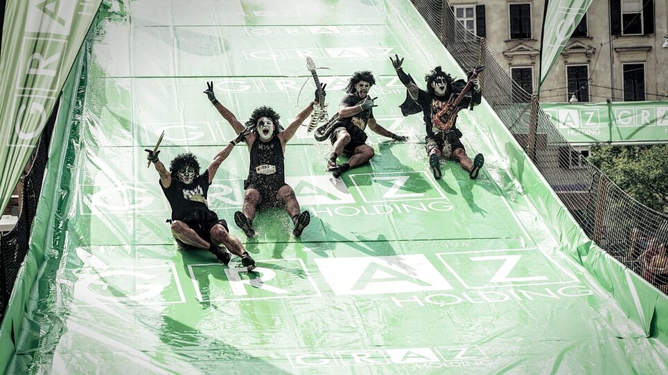 Partecipanti pazzi su uno scivolo al Grazathlon. Costumi unici e atmosfera divertente. | © SportografCompanyCode