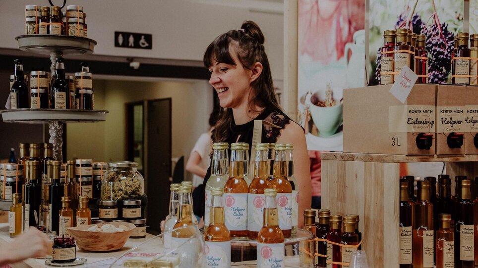 Eine fröhliche Verkäuferin präsentiert ihre Produkte auf dem Kunst- und Designmarkt in Graz. | © Kunst- und Designmarkt