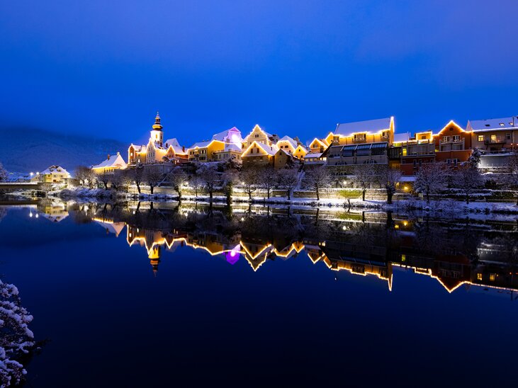 Illuminated buildings in Frohnleiten reflect in the water, surrounded by snow. | © Region Graz - Harry Schiffer