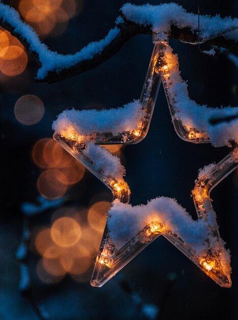 A glowing star, dusted with snow, hangs from a branch against a blurred, illuminated background. | © pixabay