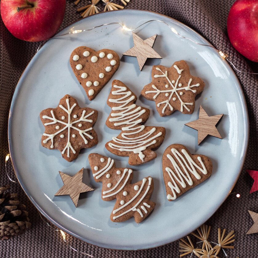 Bunte Lebkuchenformen in Herz-, Baum- und Schneeflocken-Design auf einem Teller, umgeben von Äpfeln und Holzsterne. | © pixabay