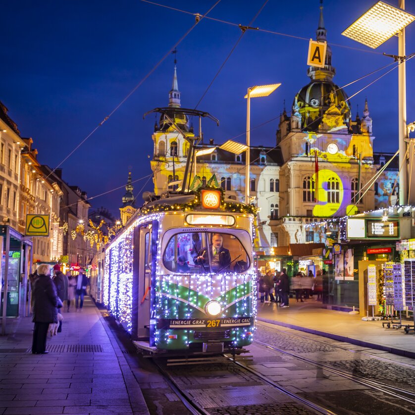 A festively lit Advent tram in Graz, surrounded by a winter scene and Christmas market. | © Graz Tourismus - Werner Krug