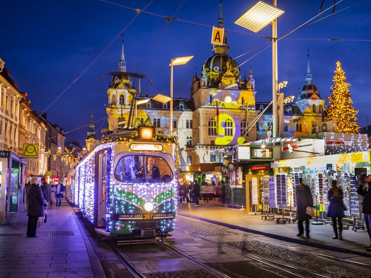 Die festlich beleuchtete Advent-Bim in Graz, umgeben von winterlicher Kulisse und Weihnachtsmarkt. | © Graz Tourismus - Werner Krug