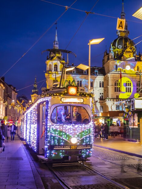 A festively lit Advent tram in Graz, surrounded by a winter scene and Christmas market. | © Graz Tourismus - Werner Krug