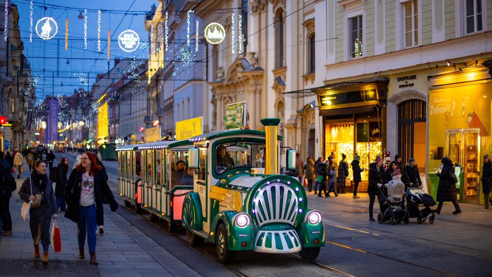 Ein festlicher Adventzug fährt durch die beleuchtete Grazer Herrengasse. Menschen schlendern entlang der Einkaufsstraße. | © Graz Tourismus - Harry Schiffer