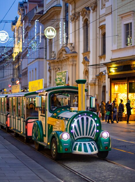 A festive Advent train passes through the illuminated streets of Graz. People stroll along the shopping street. | © Graz Tourismus - Harry Schiffer
