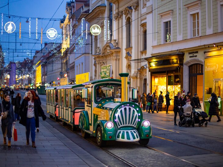 A festive Advent train passes through the illuminated streets of Graz. People stroll along the shopping street. | © Graz Tourismus - Harry Schiffer