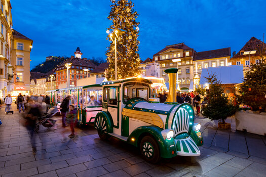 A festive city view of Graz during Advent, with a lit-up advent train driving through the streets. | © Graz Tourismus-Harry Schiffer