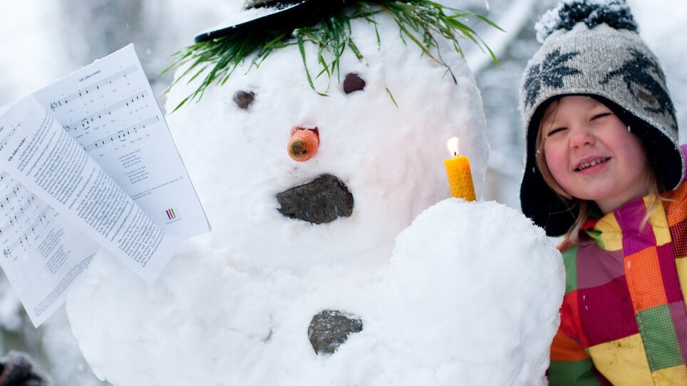 Un bambino e un pupazzo di neve con una candela e spartiti. | © Steirisches Volksliedwerk - Unterrainer