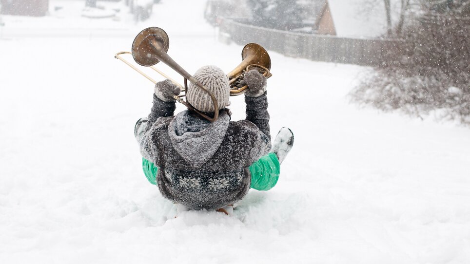 Una persona seduta su una slitta in un paesaggio innevato che suona il trombone. | © Steirisches Volksliedwerk - Unterrainer