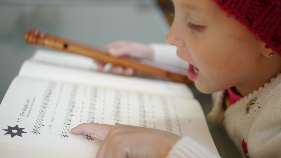 Una bambina con un berretto rosso canta e suona il flauto, guardando le note. | © Steirisches Volksliedwerk
