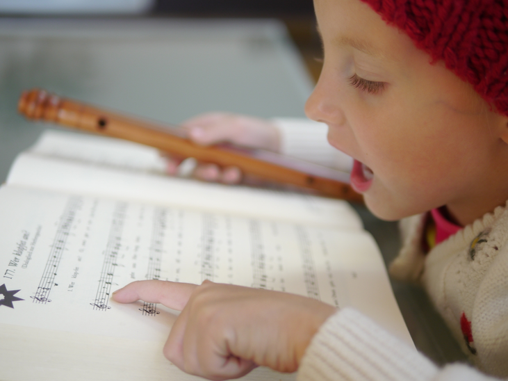 Ein Mädchen mit roter Mütze singt und spielt Flöte, während sie auf Notenblätter schaut. | © Steirisches Volksliedwerk