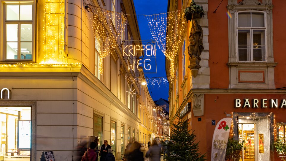 Strade illuminate con cartello 'Via dei Presepi' a Graz al crepuscolo. | © Graz Tourismus-Harry Schiffer