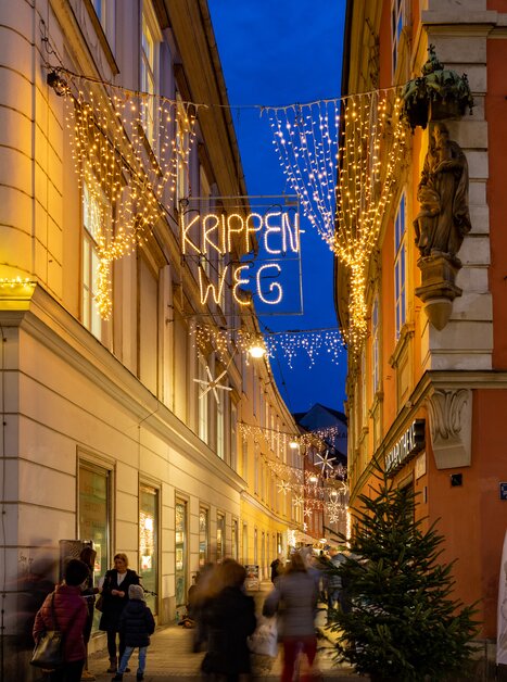 Beleuchtete Straßen mit 'Krippen Weg' Schild in Graz während der Abenddämmerung. | © Graz Tourismus-Harry Schiffer