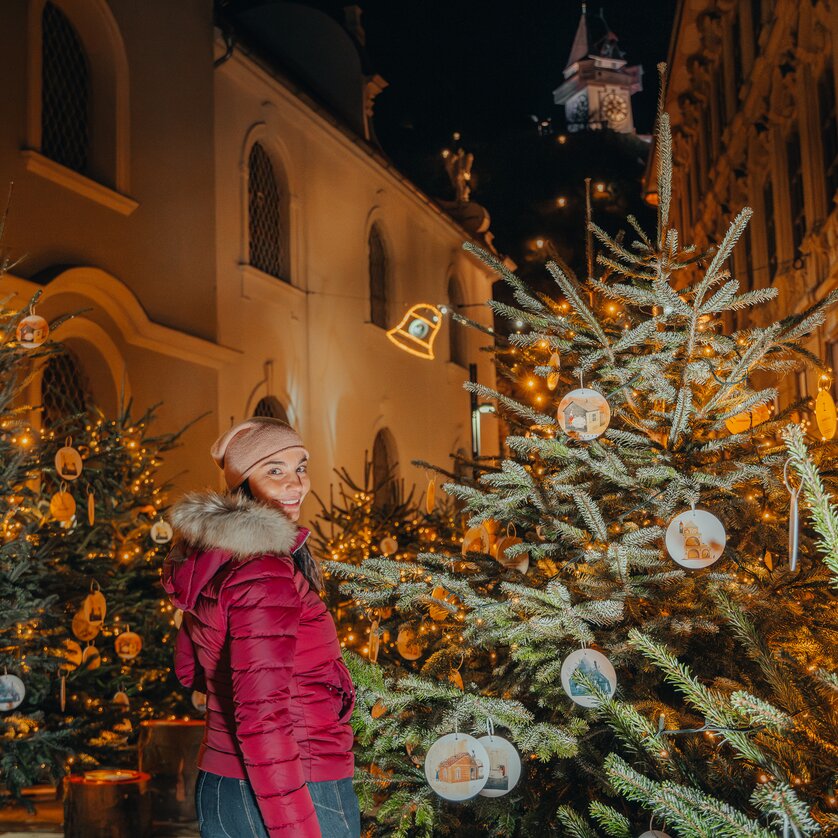 Eine Frau in einer roten Jacke steht zwischen geschmückten Weihnachtsbäumen in Graz, mit dem Uhrturm im Hintergrund. | © Mias Photoart 