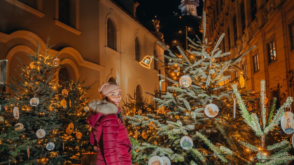 Eine Frau in einer roten Jacke steht zwischen geschmückten Weihnachtsbäumen in Graz, mit dem Uhrturm im Hintergrund. | © Mias Photoart 