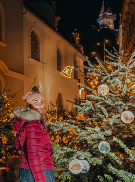 A woman in a red jacket stands among decorated Christmas trees in Graz, with the clock tower in the background. | © Mias Photoart 