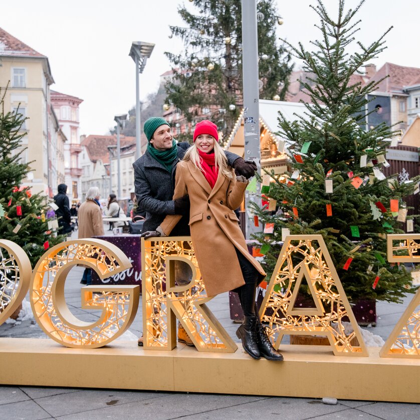 Paar posiert vor beleuchteter Graz-Dekoration im Weihnachtsmarkt. | © Graz Tourismus - Mias Photoart