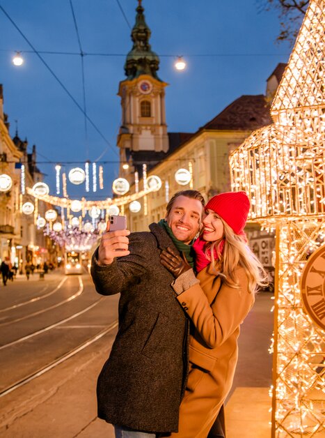 Couple takes a selfie in front of the illuminated clock tower in Graz. | © Graz Tourismus - Mias Photoart