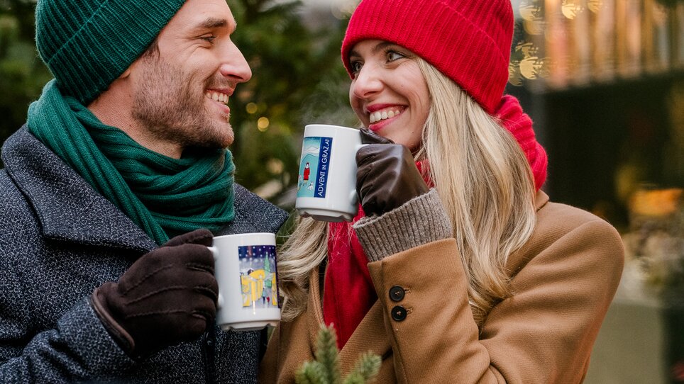 A couple with mugs in a winter atmosphere in front of a Christmas tree. | © Graz Tourismus - Mias Photoart