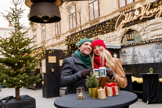 Couple in winter attire holding mugs in a festive setting. | © Graz Tourismus - Mias Photoart