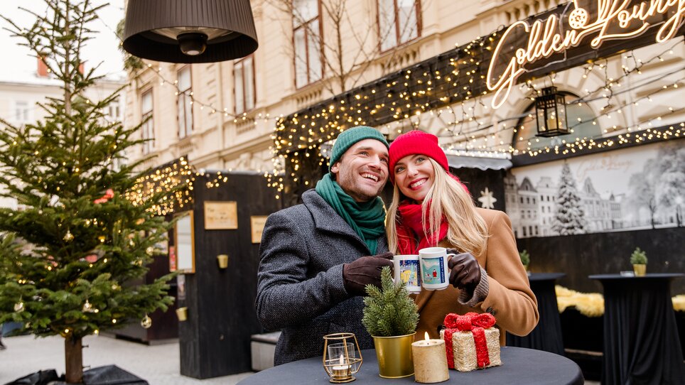 Couple in winter attire holding mugs in a festive setting. | © Graz Tourismus - Mias Photoart