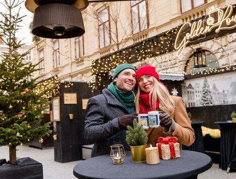 Paar in winterlicher Kleidung mit Tassen in der Hand vor festlicher Kulisse. | © Graz Tourismus - Mias Photoart