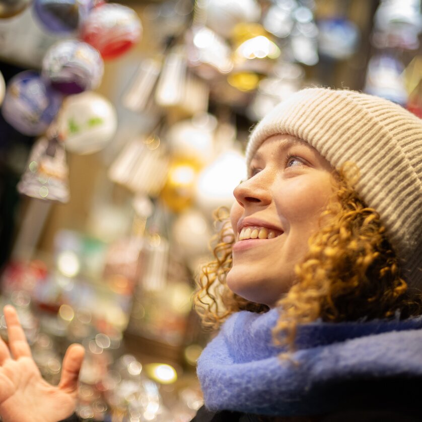 Eine fröhliche Frau mit lockigen Haaren und Mütze schaut bewundernd auf Weihnachtsdekorationen. | © Graz Tourismus - Harry Schiffer