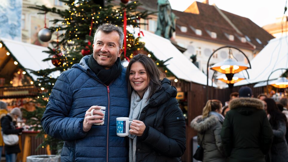 A couple smiles at a Christmas market in Graz, holding mugs in their hands. | © Graz Tourismus - Katja Stadler