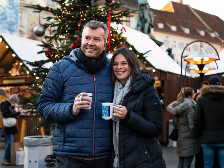 Ein Paar steht lächelnd bei einem Weihnachtsmarkt in Graz und hält Tassen in der Hand. | © Graz Tourismus - Katja Stadler