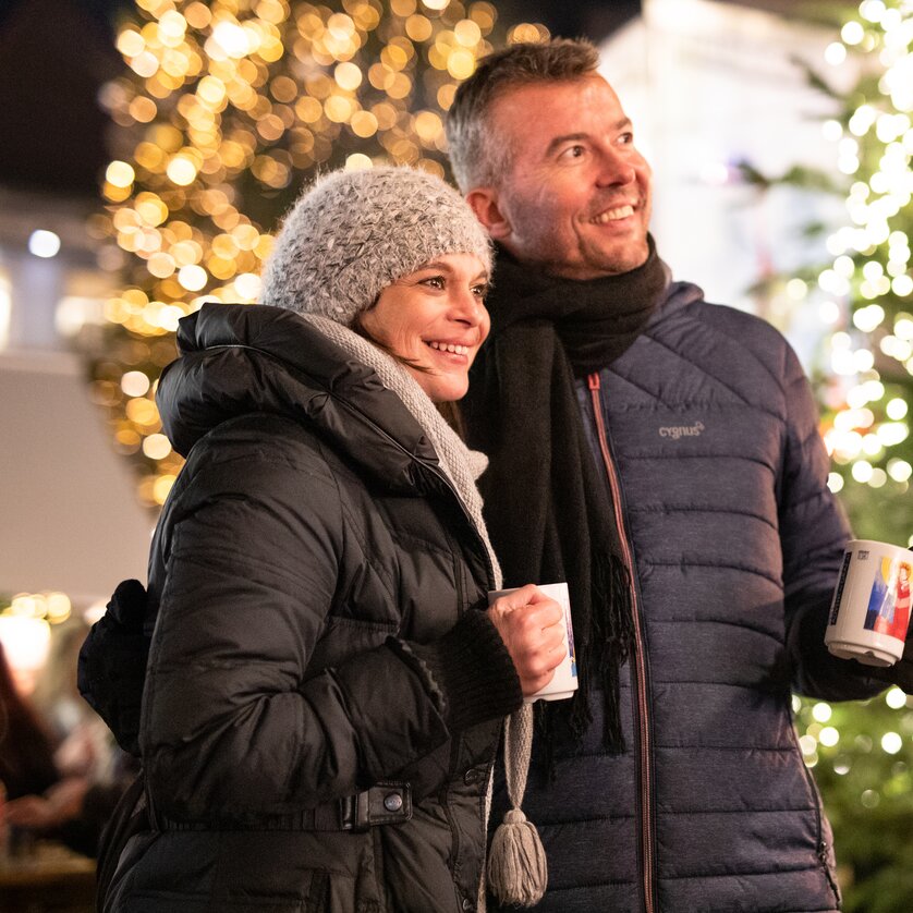 Ein Paar in Winterkleidung mit Tassen vor einem festlich beleuchteten Weihnachtsbaum. | © Graz Tourismus - Katja Stadler