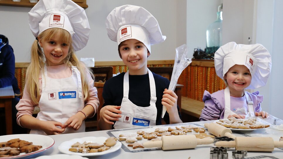 Drei Kinder mit Kochmützen backen Lebkuchen und Weihnachtskekse. | © UMJ - J. J. Kucek