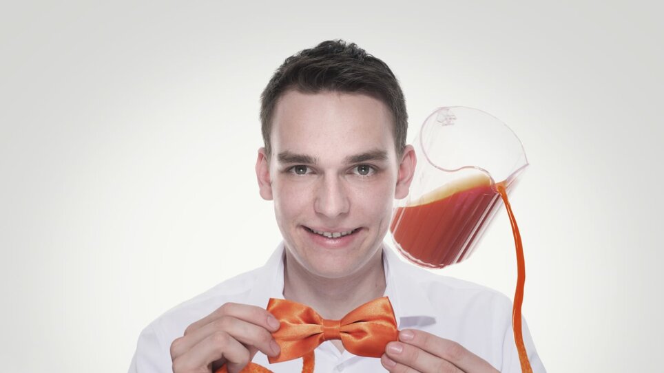 Ein Mann hält eine orangefarbene Fliege und lächelt, während ein Krug mit Saft hinter ihm schwebt. | © Zauberer Patrick