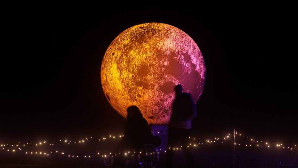 Eine große, leuchtende Mondskulptur bei Nacht mit Menschen. | © Erwin Scheriau