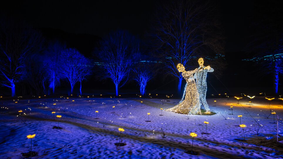Wunderschöne Lichtinstallation eines tanzenden Paares in einem Park, umgeben von beleuchteten Bäumen und einem verschneiten Boden. | © Lumagica Frohnleiten