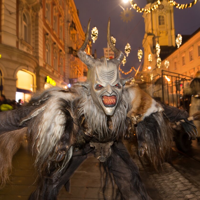 Ein Krampus mit gruseligem Gesicht und zotteliger Kleidung tanzt auf der Straße in Graz. | © Graz Tourismus - Harry Schiffer