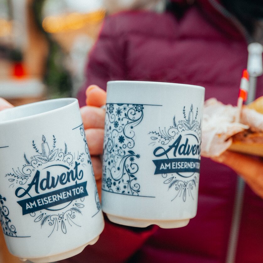 Two mugs labeled 'Advent am Eisernen Tor' and a sandwich held in hands. | © Mias Photoart