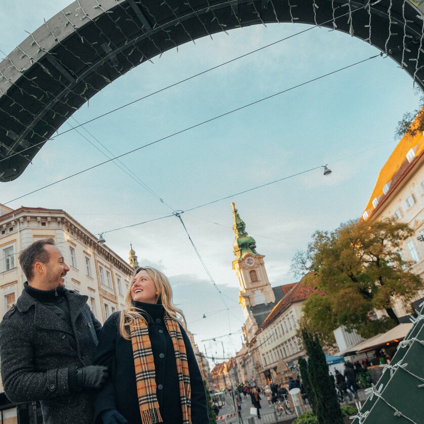 Zwei Personen im Winter auf dem Adventmarkt in Graz, mit der Stadtpfarrkirche im Hintergrund. | © Mias Photoart
