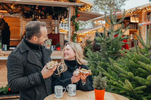 Couple at the Advent market holding snacks and cups, surrounded by lights and a Christmas tree. | © Mias Photoart