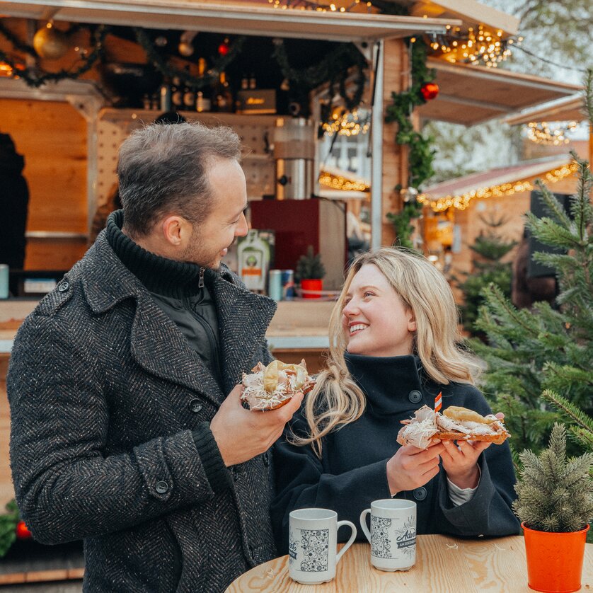 Advent in Graz I Advent market at Eisernes Tor | © Mias Photoart Couple at the Advent market holding snacks and cups, surrounded by lights and a Christmas tree. | © Mias Photoart