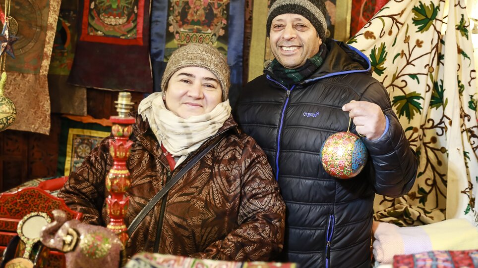 Zwei lächelnde Verkäufer stehen an einem bunten Stand mit Handwerkskunst und traditionellem Dekor. | © Thomas Luef