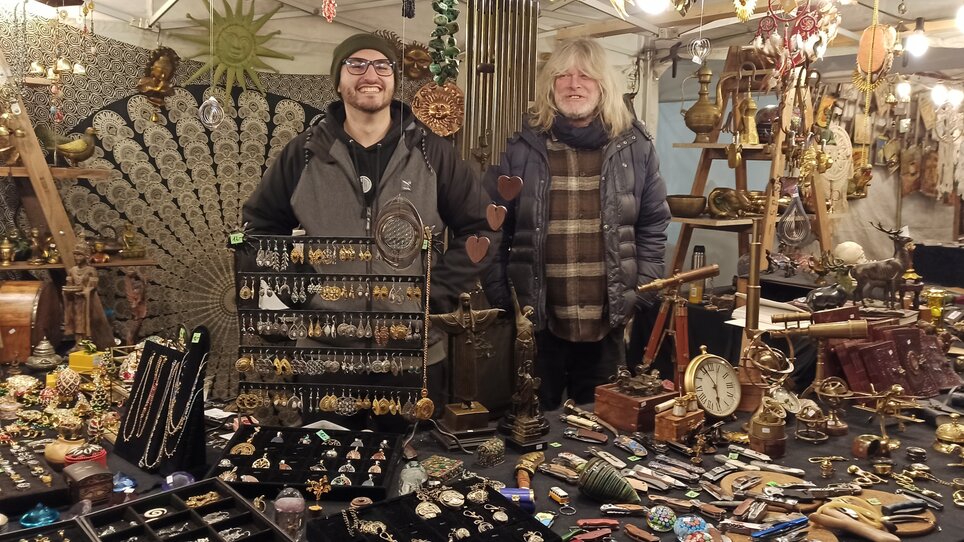 Zwei Männer stehen vor einem Tisch mit Schmuck und Kunsthandwerk auf einem Markt. | © Thomas Luef
