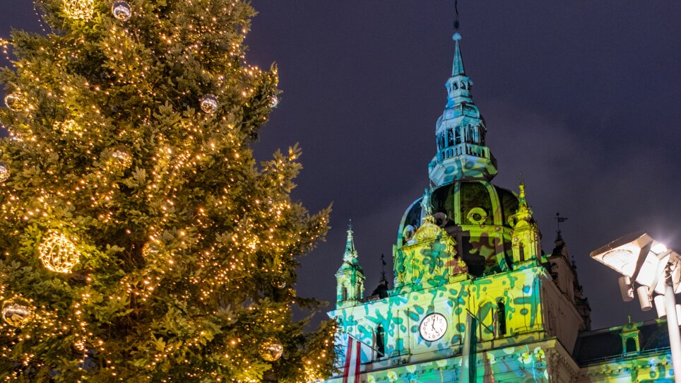 Beleuchteter Weihnachtsbaum und fröhliche Menschen vor dem Rathaus in Graz, mit festlicher Projektion auf der Fassade. | © achtzigzehn - Hinterleitner