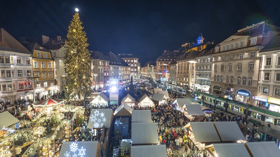 Beleuchteter Christkindlmarkt in Graz mit Menschenmengen, Weihnachtsbaum und Grazer Uhrturm im Hintergrund. | © Stadt Graz - Foto Fischer