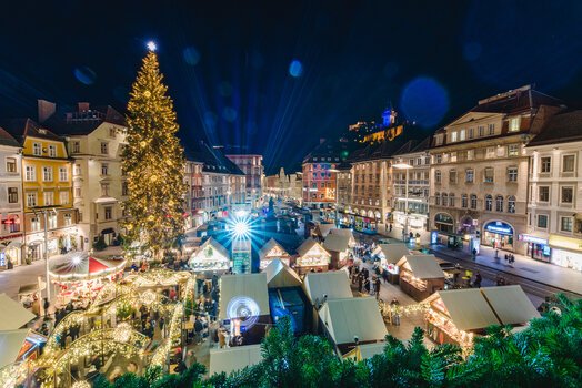 Christmas market in Graz with festively illuminated tree and Graz clock tower in the background. | © Graz Tourismus - Mias Photoart