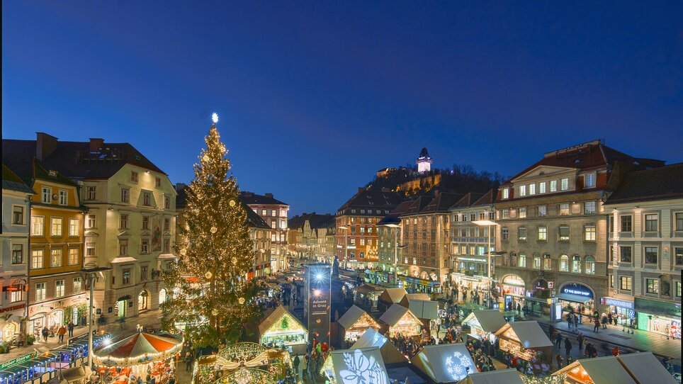 View of the Christmas market in Graz with festively decorated stalls, a large Christmas tree, and the Graz Clock Tower in the background. | © Region Graz - Robert Maybach (RBMH) 