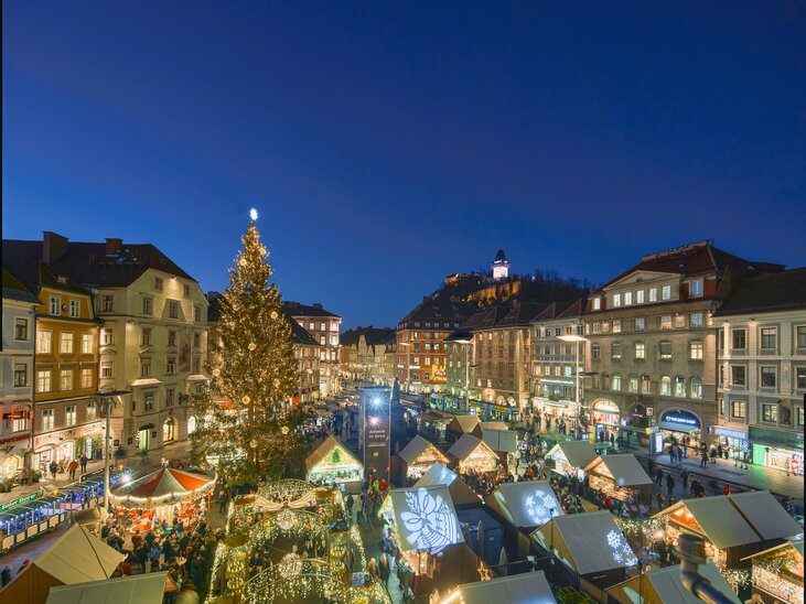 Vista del mercatino di Natale a Graz con stand decorati festivamente, un grande albero di Natale e il Torre dell'Orologio di Graz sullo sfondo. | © Region Graz - Robert Maybach (RBMH) 