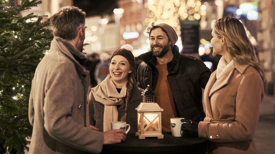 Vier Personen genießen die festliche Atmosphäre auf dem Christkindlmarkt in Graz, umgeben von beleuchteten Bäumen und einem großen Weihnachtsbaum. | © Robert Maybach