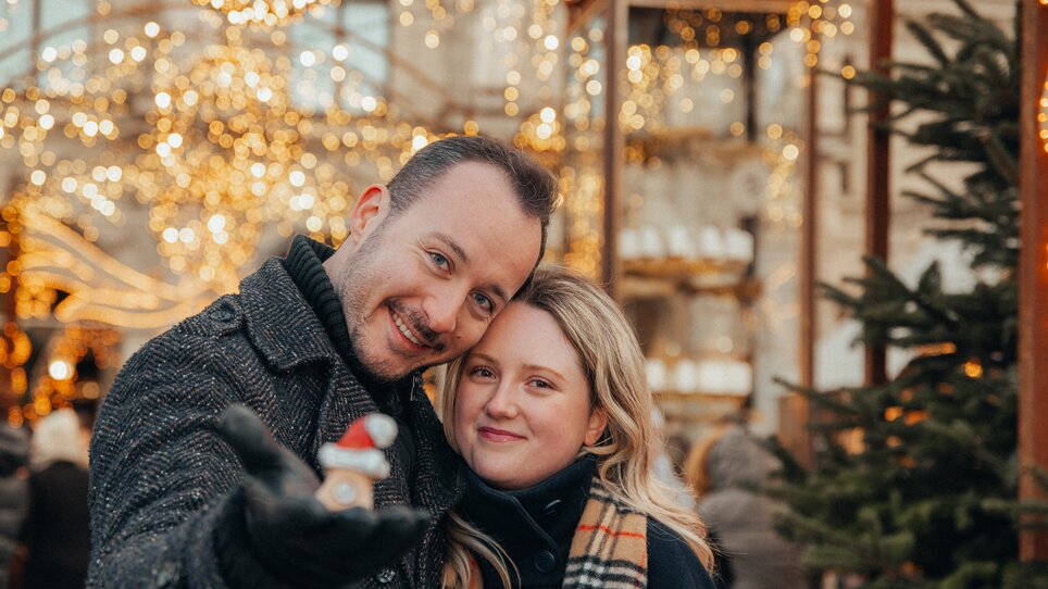 A couple poses in front of festive lights in Graz, holding Christmas decorations. | © Mias Photoart