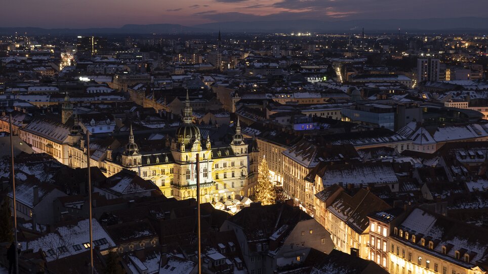 View of Graz at night with the illuminated town hall and rooftops, surrounded by snow-covered houses. | © Vincent Croce